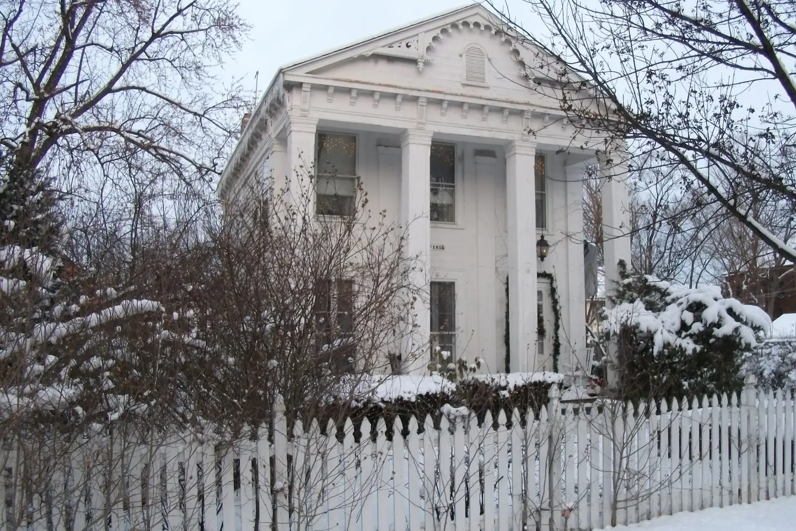Snow-covered Greek Revival house with columns behind a white picket fence in Lansingburgh